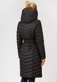 Donna con capelli biondi raccolti in un chignon che indossa un lungo piumino trapuntato nero con collo alto e vita aderente, vista da dietro.