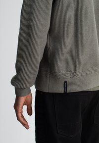 Grijze geribbelde gebreide trui met een ronde halslijn en een geweven label aan de zoom, gecombineerd met zwarte gestructureerde broek.