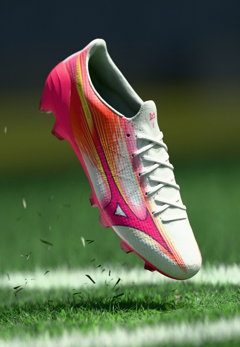 Weißer und pinker Fußballschuh mit gelben Akzenten, in der Luft über Gras auf einem markierten Fußballfeld.