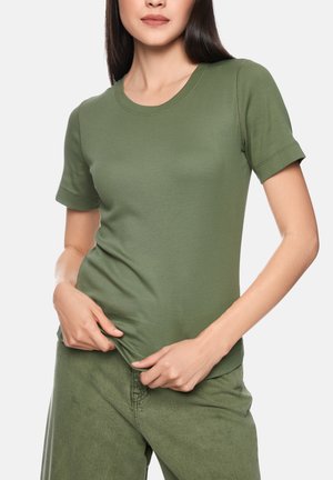 Frau trägt ein eng anliegendes olivgrünes Kurzarm-T-Shirt und passende Hosen, zieht sanft mit beiden Händen am Saum ihres T-Shirts.