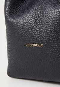 Laikku sininen nahkalaukku, jossa on teksturoitu pinta ja kultainen "Coccinelle" -logo. Muotoilussa on pehmeät, pyöristetyt reunat.