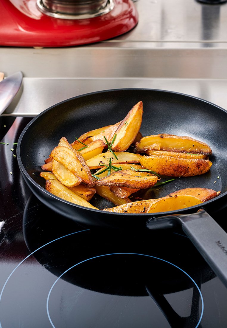 Eine schwarze Pfanne enthält goldbraune Kartoffelwedges, garniert mit grünen Kräutern, und steht auf einem glatten, schwarzen Induktionskochfeld.
