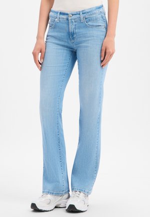 Femme portant un jean bootcut bleu clair et des baskets blanches, debout devant un fond uni.