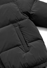 Giacca nera imbottita con trapuntatura orizzontale e una tasca verticale con zip su una superficie di tessuto liscia.