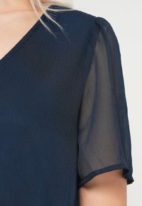 Λεπτομέρεια από μια ναυτική μπλε μπλούζα με V-λαιμό και διαφανή κοντά μανίκια σε ένα άτομο με ανοιχτόχρωμη επιδερμίδα και ξανθά μαλλιά.