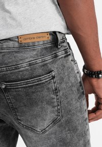 Harmaat farkut, joissa on haalistunut tekstuuri, varustettuna nahkapaikalla, jossa lukee "ombre denim", sekä kahdella takataskulla metallikoristeilla.