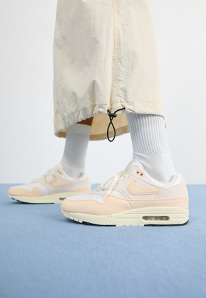 A Nike Air Max 1 könnyű, fehér és puha barack színű felsőrésszel rendelkezik, amely hálós és bőr anyagokból készült, látható légpárnával és textúrázott talppal.