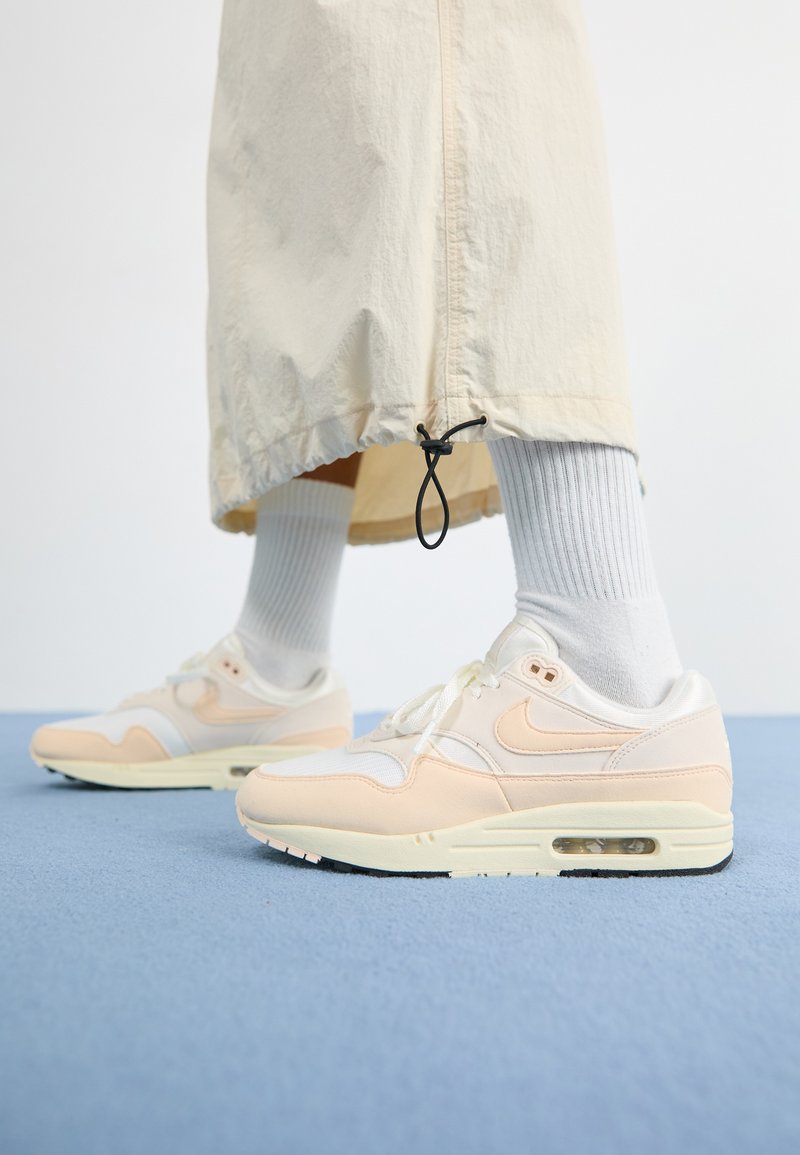 Les baskets Nike Air Max 1 légères présentent un dessus blanc et pêche doux en matériaux mesh et cuir, une unité Air visible et une semelle texturée.