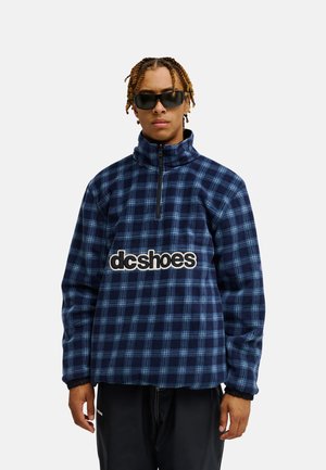 Mann mit schwarzen Sonnenbrillen und einem blau karierten Fleece-Pullover mit "dc shoes" Logo auf der Brust, steht vor einem einfarbigen hellen Hintergrund.