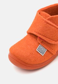 Oranžová plstená obuv s okrúhlym špičkou, vybavená suchým zipsom a gumovou podrážkou. Obsahuje nášivku s logom na boku.