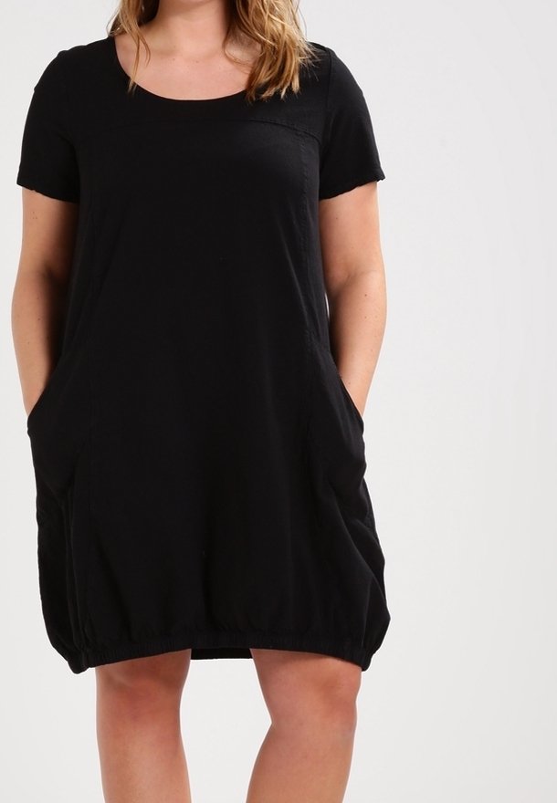 Femme portant une robe noire à manches courtes avec un col rond et des poches, debout devant un fond clair uni.