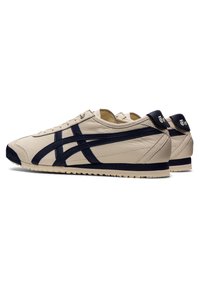 Beige leather sneakers with navy blue stripes, rounded toe, flat rubber sole, and contrasting navy heel tab. Classic design with subtle texture.