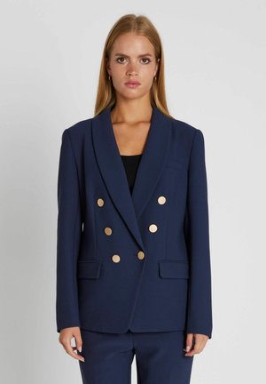 Młoda kobieta ubrana w granatową marynarkę dwurzędową z złotymi guzikami i pasujące spodnie, stojąca na jasnym, jednolitym tle.