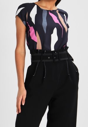 Femme portant un chemisier noir à manches courtes avec des formes abstraites multicolores et un pantalon noir taille haute avec une large ceinture et des coutures contrastées.