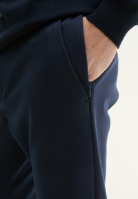 Des joggings bleu marine en tissu doux présentent une poche latérale avec fermeture éclair. La taille est élastique, offrant un ajustement confortable.