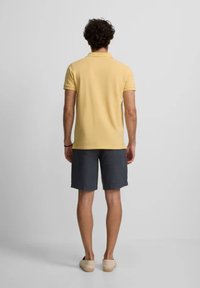 Camisa polo de manga corta amarilla, pantalones cortos grises y zapatos de color claro. Vista posterior, tela suave y diseño informal.