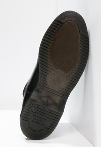 Suela de zapato negra con goma texturizada, presenta surcos profundos y un logo en relieve. Las marcas indican desgaste. La forma es redondeada, con una parte inferior plana.