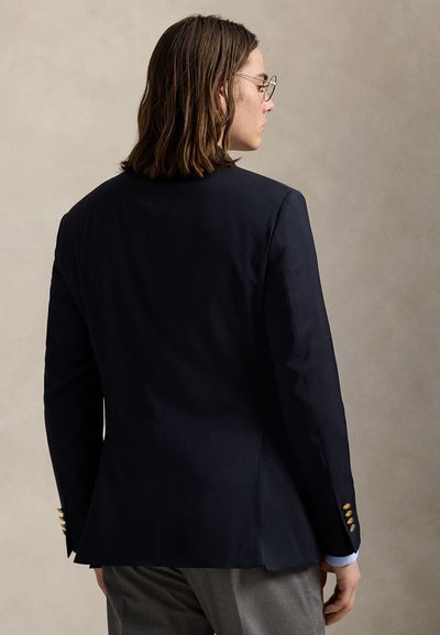 Mand med skulderlangt hår og briller, iført navy blazer med guldknapper og grå bukser, set fra siden mod beige baggrund.
