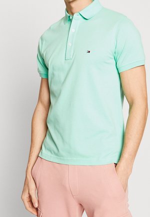 Man draagt een lichtgroen poloshirt met korte mouwen en een klein logo op de borst, en lichtroze broek met zakken.