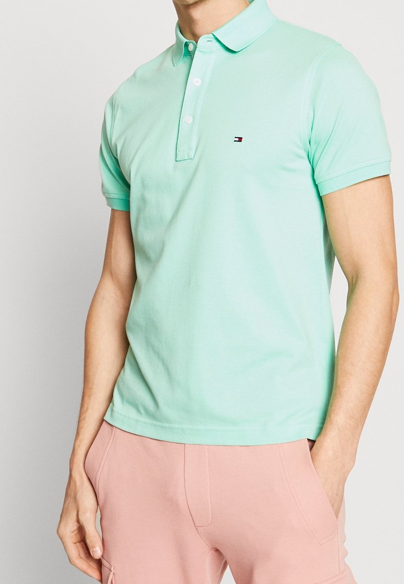 Homme portant un polo à manches courtes vert clair avec un petit logo sur la poitrine et un pantalon rose clair avec des poches.