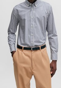 Randig skjorta med knappar och långa ärmar i marinblått och vitt, kombinerad med khakifärgade byxor och ett svart bälte. Har klassisk krage och fickor.