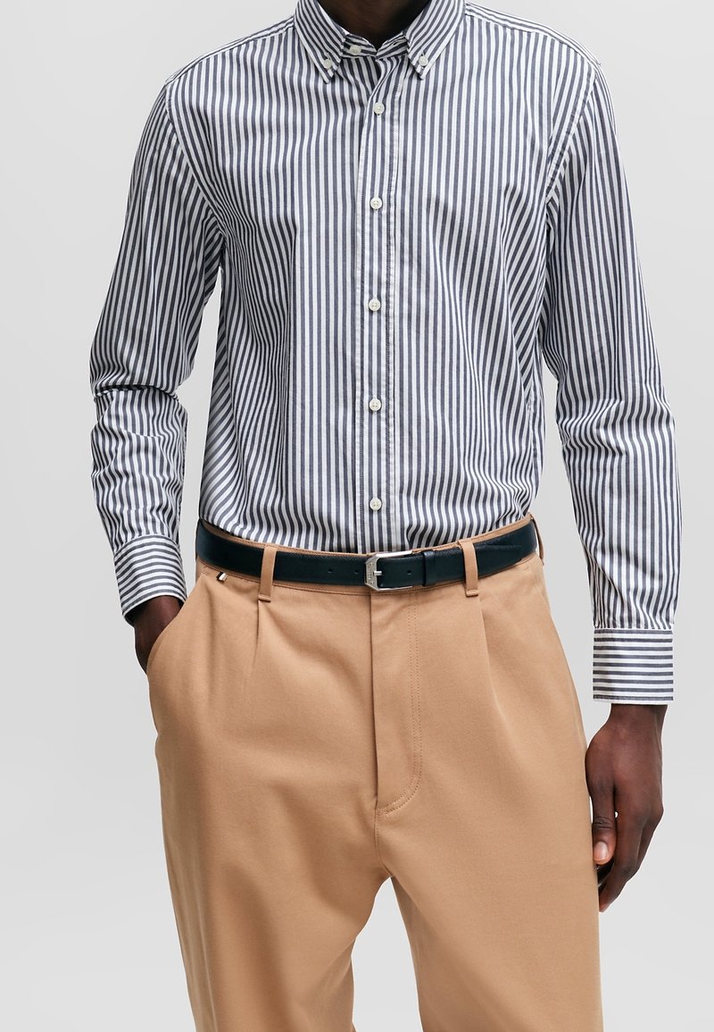 Randig skjorta med knappar och långa ärmar i marinblått och vitt, kombinerad med khakifärgade byxor och ett svart bälte. Har klassisk krage och fickor.