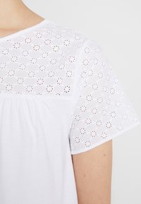 Biała koszula z teksturowanym wzorem ażurowym na rękawach, z gładkim przodem i okrągłym dekoltem. Luźny krój z marszczeniem.