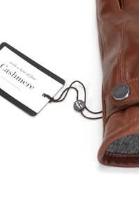 Ruda odinė pirštinė su pilku kašmyro pamušalu, suapvalintu metaliniu mygtuku ir etiketė, pritvirtinta tamsia virvele.