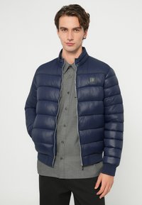 Tengerészkék steppelt mintájú tollkabát, cipzáras lezárással és magas gallérral. Szürke, gombos ing fölött viselve, a mellrészen logóval ellátott folttal.