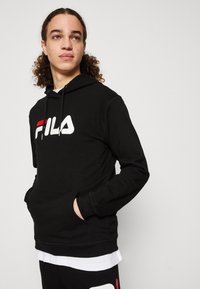 Felpa nera con cappuccio e tasca frontale, adornata con un logo FILA bianco e rosso. Tessuto liscio e polsini a costine, sovrapposta a una maglietta bianca.