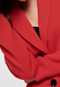 Blazer rojo con un diseño estructurado, mangas largas y cierre de un solo botón. Tejido suave con un acabado ligeramente texturizado.