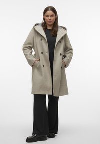 Beige uldfrakke med hætte, dobbeltradet design, store knapper og sidelommer. Båret over en sort top og brede sorte bukser.