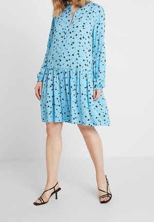 Mujer con un vestido suelto de manga larga azul claro con estampado floral negro y sandalias negras de tacón con tiras, posando frente a un fondo blanco.