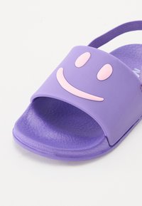 Lilla gummisandaler med rutsjebane-design og struktureret fodseng, der har en pink smiley ansigt på remmen og en baghælsrem for støtte.