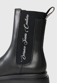 Black Chelsea boot featuring a smooth leather finish, elastic side panels, and embossed "Versace Jeans Couture" branding. Thick sole.