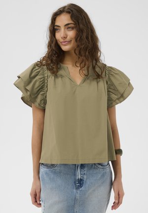 Kobieta o kręconych włosach nosi oliwkowo-zielony top z krótkim rękawem i falbanami na ramionach oraz jasnoniebieską spódnicę dżinsową na jednolitym tle.