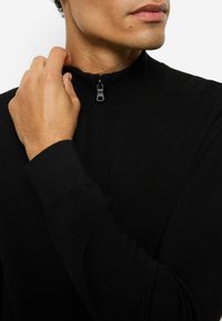 Schwarzer, strukturierter Reißverschluss-Pullover mit hohem Kragen und kleinem Reißverschluss-Detail. Eng anliegend mit gerippten Bündchen und glattem Finish.