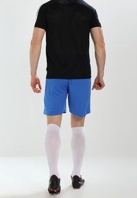 Sportovní oblečení zahrnující černé tričko s krátkým rukávem, modré šortky, bílé vysoké ponožky a černé kopačky, z pohledu zezadu.