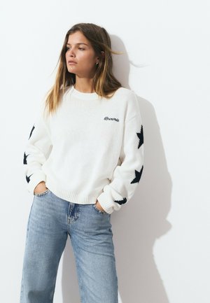 White knitted sweater with black star patterns on sleeves; features "Brownie" logo. Paired with light blue jeans, showcasing relaxed fit.