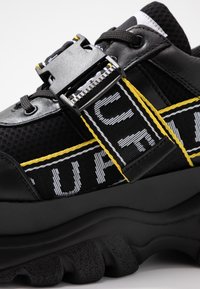 Svart atletisk sko med mesh- och syntetmaterial, gula och vita accentränder, spänne och en strukturerad chunky sula.