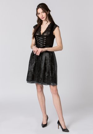 Junge Frau steht, trägt ein ärmelloses schwarzes Spitzenkleid mit Schleifendetail und schwarze High Heels, vor einfachem hellgrauem Hintergrund.