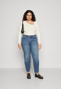 White long-sleeve top with a cutout, high-waisted blue jeans, and black loafers. Model holds a small black bag. Neutral background.