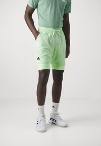 T-shirt de sport verte, shorts vert menthe avec logo Adidas noir, et chaussures blanches avec accents noirs. Ceinture élastique et cordon de serrage visibles.