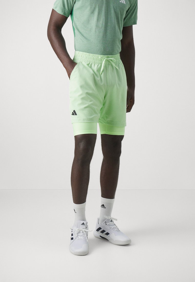 T-shirt de sport verte, shorts vert menthe avec logo Adidas noir, et chaussures blanches avec accents noirs. Ceinture élastique et cordon de serrage visibles.