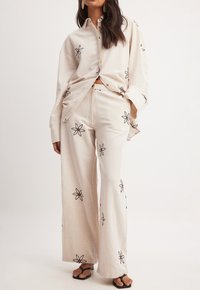 Beige linnen overhemd en wijde broek met zwarte bloemige borduring. Het overhemd heeft een knoopsluiting en lange mouwen.