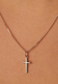 Zilveren kruis hanger aan een dunne, gedraaide ketting, rustend tegen een huidkleurige achtergrond, die het gladde, gepolijste oppervlak benadrukt.