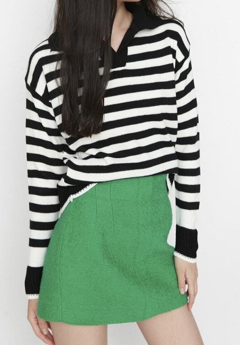 Femme portant un pull à rayures horizontales noires et blanches rentré dans une mini-jupe verte vive texturée, debout devant un fond blanc.