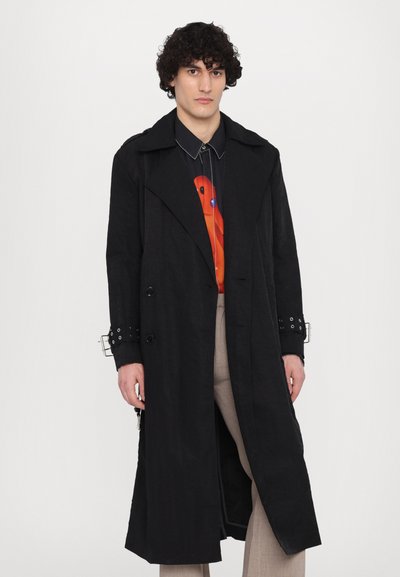 Schwarzer, langer Trenchcoat mit doppelt geknöpften Knöpfen, breiten Revers und gestanzten Manschetten, kombiniert mit einem bunten Hemd und hellen Hosen.
