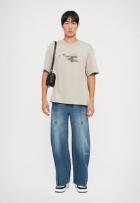Beige bomull t-skjorte med grafisk design, løs passform. Bred ben blå denim jeans med blekede seksjoner. Svart crossover-veske, hvite sneakers.