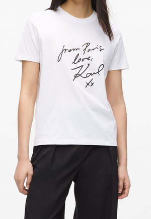 Kobieta ubrana w białą koszulkę z czarnym, odręcznym napisem "from Paris love, Karl xx" i czarne spodnie na jednolitym tle.